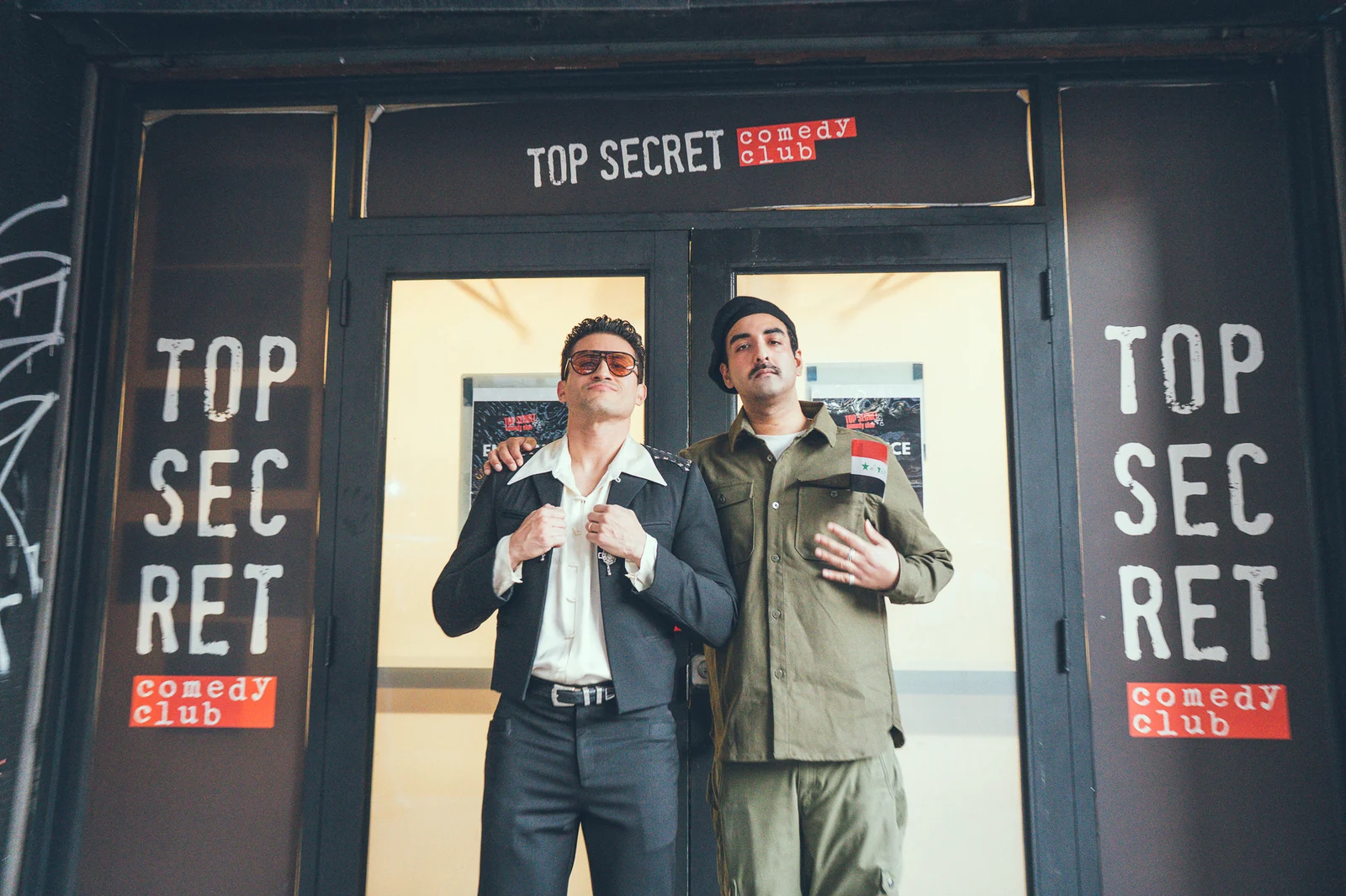 Two people in performance outfits pose together outside the Top Secret Comedy Club entrance.