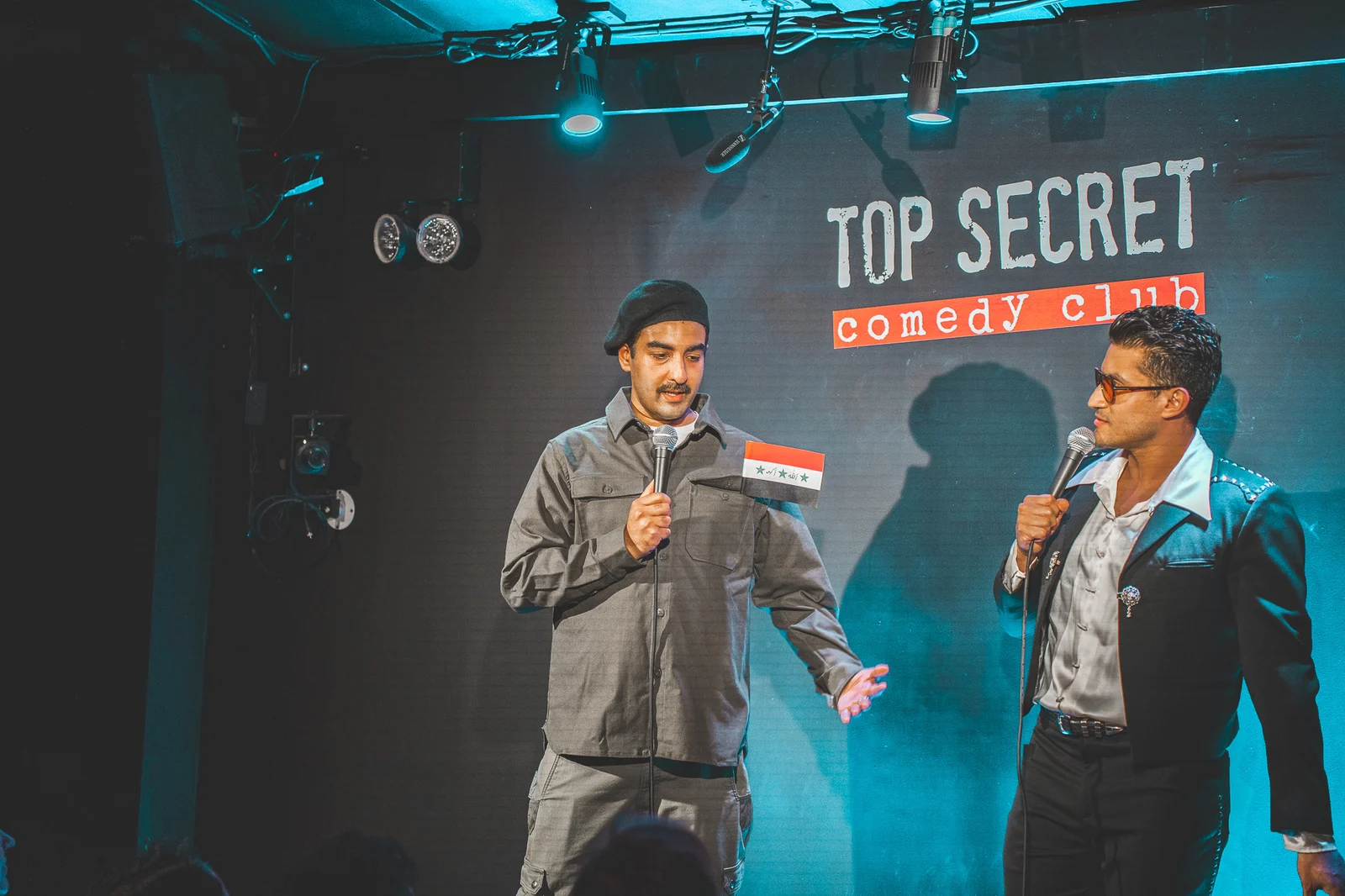 Two performers hold microphones, one extending an open hand, beneath the Top Secret Comedy Club sign.