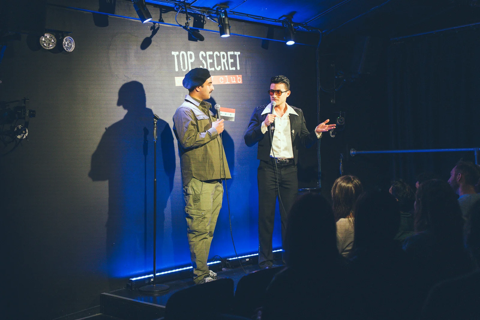 Two performers exchange lines with microphones on a blue-lit stage, with the audience in the foreground.